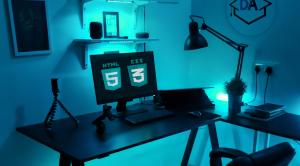 Blue lit at home office with html 5 and css 3 logo on computer screen and debug academy logo as a poster on the wall